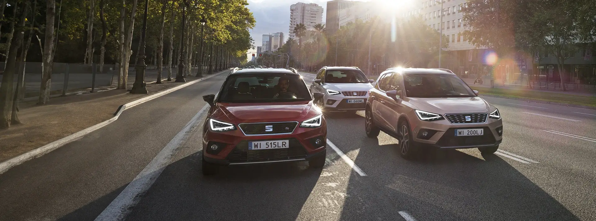 trzy SUV-y SEAT Ateca w kolorach czerwonym, szarym i biały, jadą dwoma pasami na drodze dzielącej miasto i las