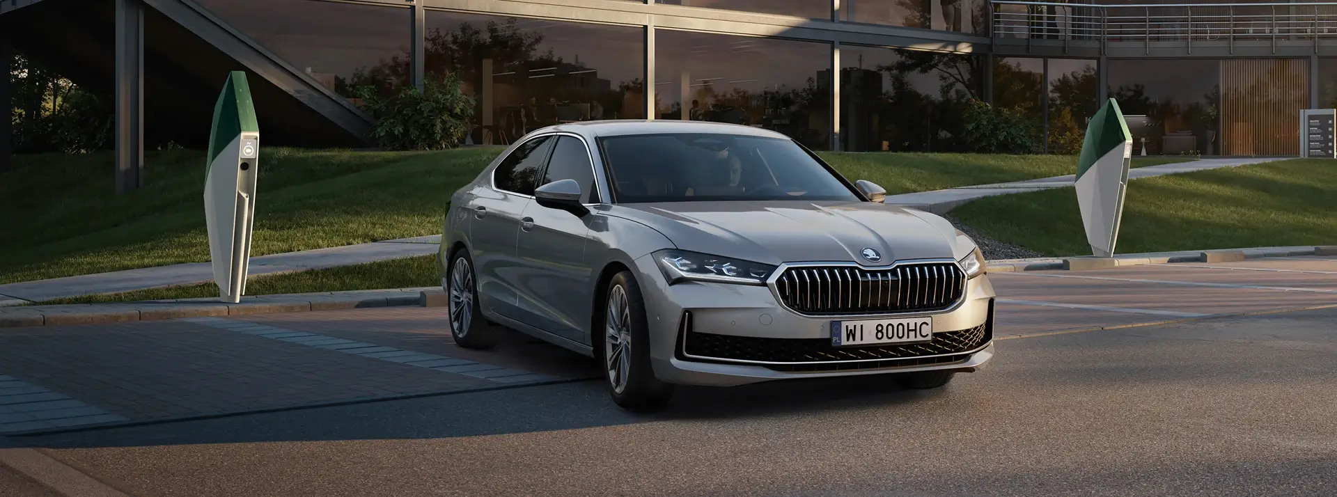 parking na tle trawy i nowoczesnego budynku, kierowca szarej limuzyny Skoda Superb właśnie parkuje