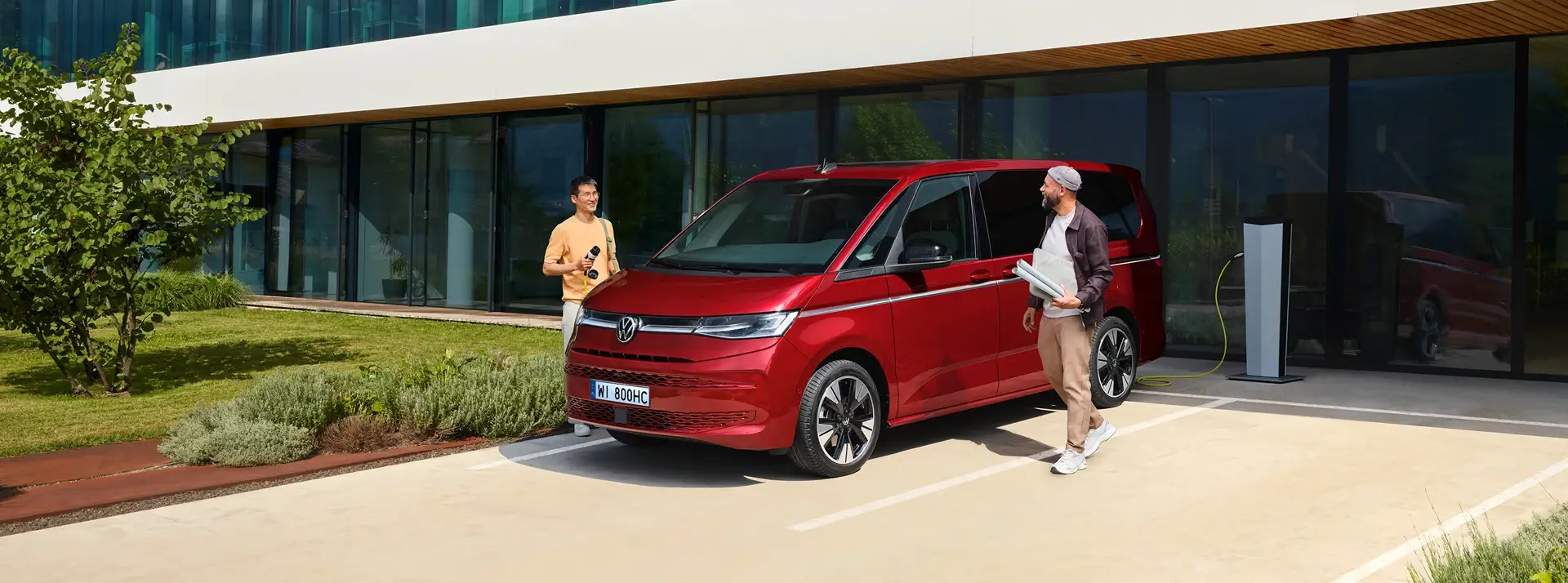 dwóch mężczyzn obok czerwonego Volkswagena Caravelle zaparkowannego przed oszklonym budynkiem